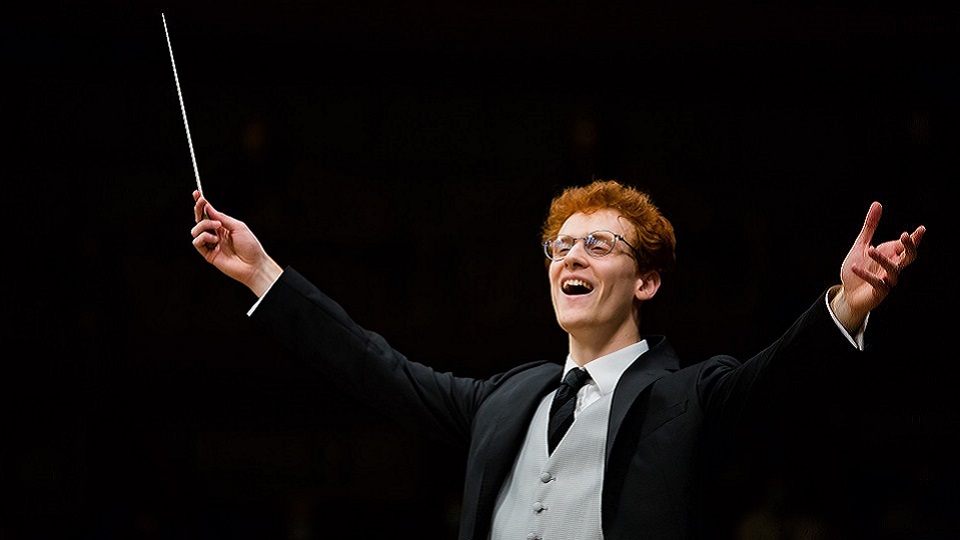 Photo of male alumnus conducting an orchestra, wearing a smart outfit and glasses